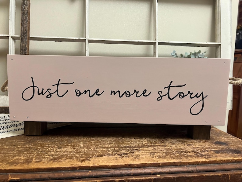 May include: A rectangular, light pink wooden box with the phrase "Just one more story" in black script. The box has dark brown wooden feet and rope handles. The background includes a window.
