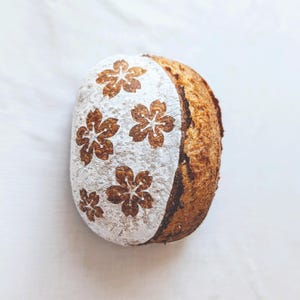 May include: A round loaf of sourdough bread with a golden-brown crust. One side is dusted with white flour and features a decorative pattern of five cherry blossom flowers scored into the surface, revealing the dark brown dough underneath.