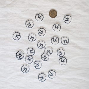 Puede incluir: Un conjunto de pequeños parches circulares blancos con un diseño de 'n' negro, dispersos sobre una tela blanca. También hay una moneda. Los parches parecen hechos a mano, posiblemente para manualidades o adornos.