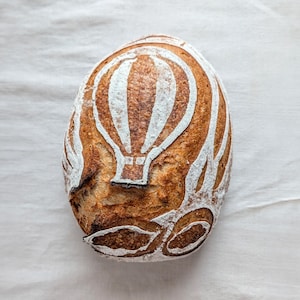 May include: A round loaf of sourdough bread with a golden-brown crust. The top surface features a decorative score pattern of a hot air balloon, created by dusting white flour into the carved lines against the dark, baked bread surface.