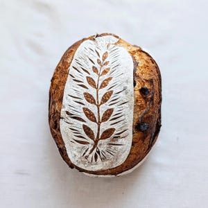 May include: A round loaf of sourdough bread with a dark brown, crispy crust. The top surface features a decorative wheat stalk pattern scored into the dough, highlighted by a dusting of white flour that contrasts with the baked golden-brown color.