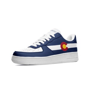 May include: White and navy blue sneakers with a white stripe and the Colorado state flag on the side. The flag is red, yellow, and blue.