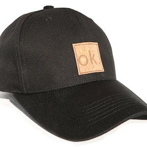 Könnte beinhalten: Eine schwarze Baseballkappe mit einem braunen Kork-Patch, auf dem die Buchstaben "OK" in einem Quadrat stehen.