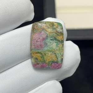 Pode incluir: Um cabochão de pedra preciosa polida de formato retangular com um padrão marmorizado de inclusões em magenta vibrante, verde musgo e amarelo dourado. A pedra tem uma superfície lisa e brilhante com texturas minerais naturais visíveis.