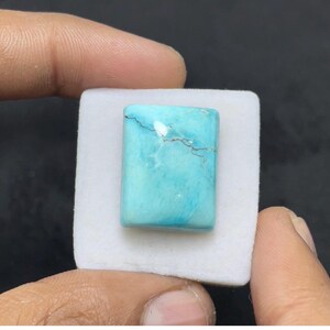 Puede incluir: Una piedra preciosa de turquesa pulida de forma rectangular, con un vibrante color azul cielo, un sutil veteado blanco y una fina veta de matriz marrón oscuro que atraviesa horizontalmente la parte superior, montada sobre una pequeña almohadilla de espuma blanca.