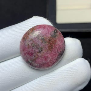 Puede incluir: Un cabujón de piedra preciosa redondo y pulido que presenta un color base magenta vibrante y rosa intenso con inclusiones minerales de color gris oscuro y marrón dispersas por toda su superficie cristalina texturizada y acabado brillante.