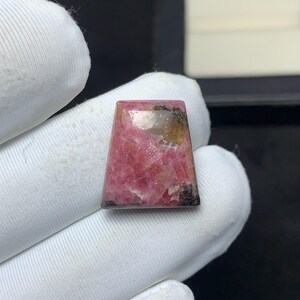Può includere: Un cabochon di rodocrosite trapezoidale con una superficie lucida caratterizzata da un vivace motivo cristallino rosa e bianco, accentuato da inclusioni minerali scure sui bordi superiore e inferiore, con una finitura liscia e riflettente.