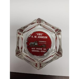 May include: Clear, hexagonal ashtray with a red and white circular graphic. The graphic features the text "VIRG" and "V. W. JOHNSON", along with a vintage car illustration and the text "CONOCO". Additional text includes "1653 PHONE 213 GRINNELL, IOWA".