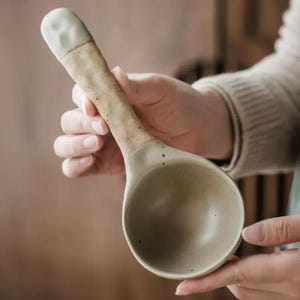 May include: A ceramic serving spoon with a long handle and a rounded bowl. The handle is a speckled tan colour, transitioning to a light green-grey bowl. The tip of the handle is a solid, light green-grey colour.