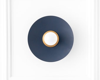 May include: A navy blue wall sconce with a circular design. The sconce features a dark blue outer ring, a gold inner ring, and a white center light. The fixture is mounted on a white wall.