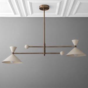 May include: A linear chandelier featuring a central bronze-toned rod and horizontal arms. Two off-white conical shades are positioned at opposite ends, each containing a light bulb. Two small spherical accents are attached to the horizontal arms.
