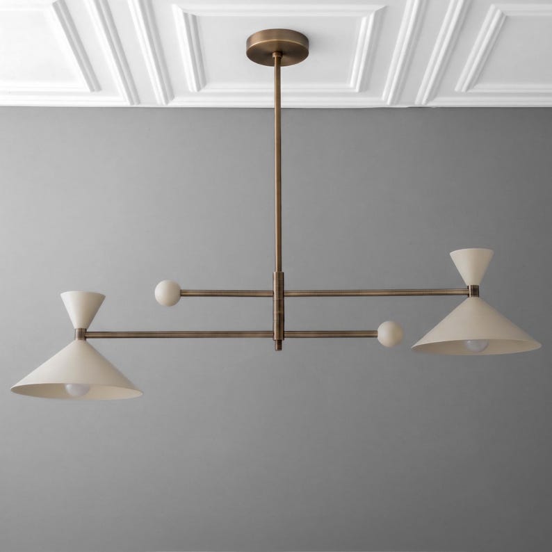May include: A linear chandelier featuring a central bronze-toned rod and horizontal arms. Two off-white conical shades are positioned at opposite ends, each containing a light bulb. Two small spherical accents are attached to the horizontal arms.
