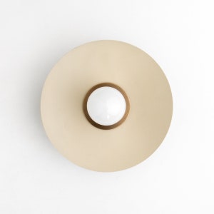May include: A modern, circular wall sconce with a cream-colored disc and a central, white, spherical light. The light is surrounded by a bronze-colored ring. The sconce is mounted on a white wall.
