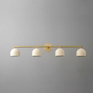 May include: A four-light vanity fixture with a gold finish. The fixture features four off-white, dome-shaped shades. The shades are evenly spaced along a horizontal bar, attached to a circular wall mount.