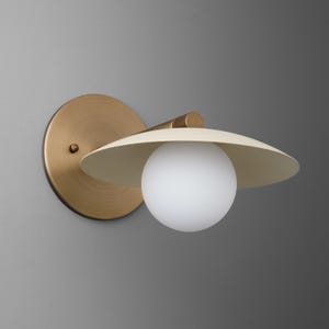 May include: A modern wall sconce with a brass finish. The sconce features a circular backplate, a curved arm, and a cream-colored, hat-shaped shade. A white globe-shaped light bulb is centered beneath the shade. The fixture is mounted on a gray wall.