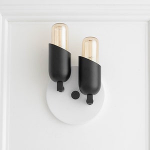May include: A black wall sconce with two light bulbs. The sconce has a white circular base and is mounted on a white wall.