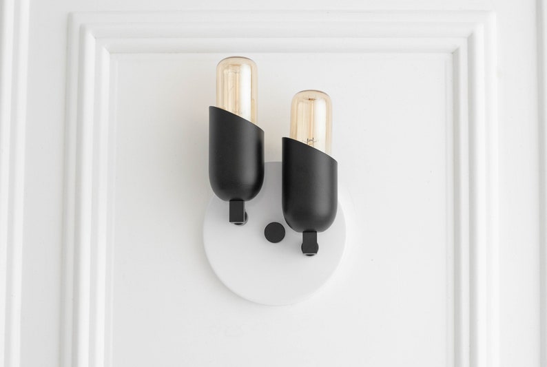 May include: A black wall sconce with two light bulbs. The sconce has a white circular base and is mounted on a white wall.