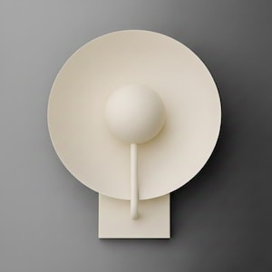 May include: Cream-colored wall sconce with a circular shade and a spherical accent. The sconce has a stem-like support and a square base. The design is minimalist and modern, with a matte finish. The sconce is mounted on a gray wall.