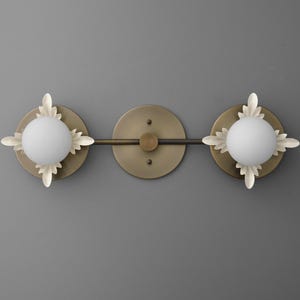 May include: A wall-mounted light fixture featuring a horizontal bronze-toned bar with a central circular plate and two spherical white glass globes. Each globe is surrounded by a decorative off-white metal leaf-shaped frame.