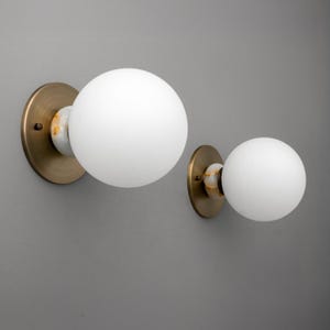 May include: Two wall sconces with a brass finish and white globe shades. The sconces have a marble accent between the brass base and the globe. The sconces are mounted on a gray wall.