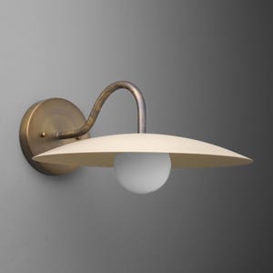 May include: A wall-mounted light fixture with a brass-colored circular base and curved arm. The fixture features a cream-colored, saucer-shaped shade and a white globe light. The design is minimalist and retro.