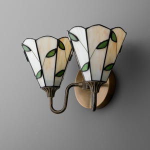 May include: A two-light wall sconce featuring stained glass shades in a cream and white hue with green leaf accents. The shades are shaped like inverted flowers and are supported by a circular bronze-toned metal base with curved arms.