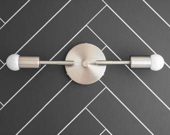 May include: A brushed nickel wall sconce with two frosted glass bulbs. The sconce is mounted on a black tiled wall.