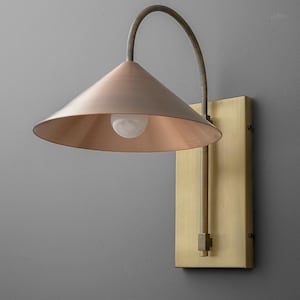 May include: A wall-mounted light fixture with a copper-colored conical shade and a brass-colored rectangular backplate. The fixture has a curved arm and a visible light bulb. The design is industrial and modern.