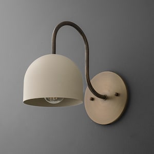 May include: A wall-mounted light fixture with a cream-colored, dome-shaped shade and a dark brown, curved arm. The fixture is attached to a round, brass-colored base. The light bulb is visible inside the shade.