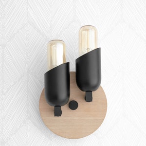 May include: A pair of black wall sconces with round wooden bases and clear glass bulbs. The sconces are mounted on a white wall.