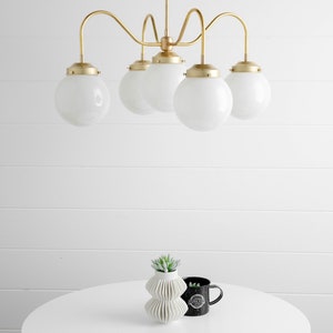 May include: A gold five-light chandelier with white globe-shaped glass shades hangs from the ceiling above a white table with a small green plant in a white vase and a black mug.