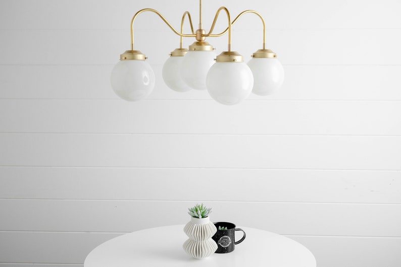 May include: A gold five-light chandelier with white globe-shaped glass shades hangs from the ceiling above a white table with a small green plant in a white vase and a black mug.