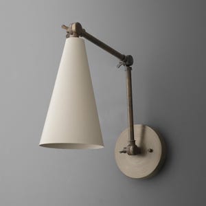 May include: A wall-mounted lamp with a cream-colored, cone-shaped shade and an adjustable arm. The lamp has a circular base and a bronze finish, mounted on a gray wall. The design is minimalist and industrial.