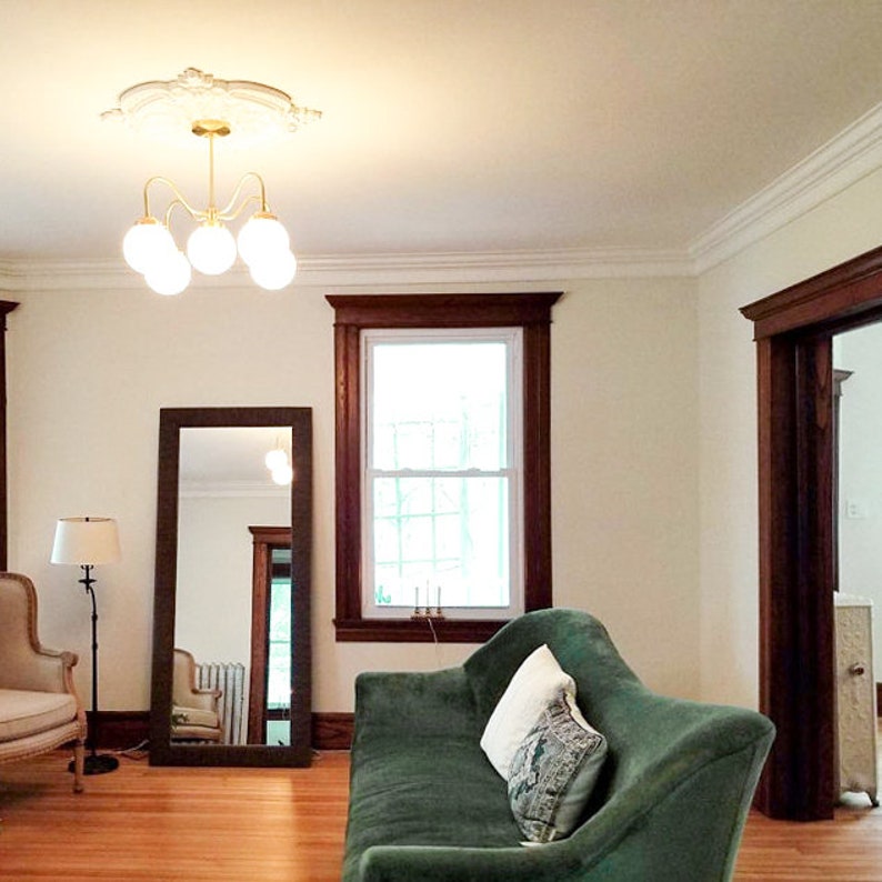 May include: A living room interior with a green velvet sofa, a full-length mirror, a lamp, and a chandelier with white globes. The room has white walls and dark wood trim around the windows and doors.