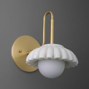 May include: A wall-mounted light fixture with a gold-colored metal base and arm. The light features a white, scalloped shade and a round, frosted glass bulb. The fixture is designed for indoor use.