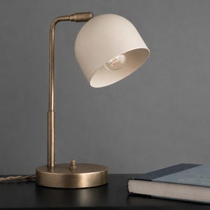 May include: A table lamp with a matte cream-colored dome shade and a metallic bronze-toned base and arm. The lamp features a slender vertical stem, a circular base with a small switch, and a fabric-covered cord extending to the left side.
