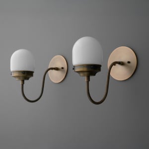May include: Two wall sconces with a vintage aesthetic. Each sconce features a white, globe-shaped glass shade, a curved, dark metal arm, and a round, brass-colored wall plate. The design evokes a classic, industrial style.