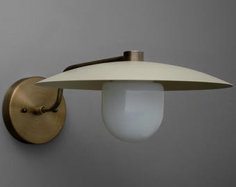 May include: A wall-mounted light fixture with a cream-colored, saucer-shaped shade and a white, rounded glass bulb. The fixture has a brass-colored circular base and a curved arm. The design is minimalist and modern.