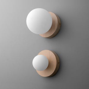 May include: Two wall sconces with a modern design. Each sconce features a round, light-colored wooden base with a white, spherical light fixture. The sconces are mounted on a gray wall, creating a minimalist aesthetic.