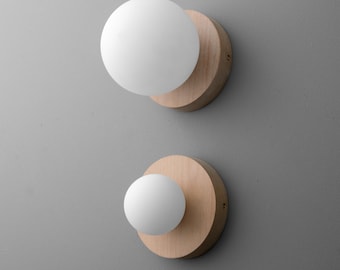 May include: Two wall sconces with a modern design. Each sconce features a round, light-colored wooden base with a white, spherical light fixture. The sconces are mounted on a gray wall, creating a minimalist aesthetic.