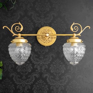 May include: A gold-toned wall light fixture featuring two clear, textured glass shades with a bubble pattern. The fixture has a central circular mounting plate with an ornate embossed design and two decorative scroll arms extending to each side.