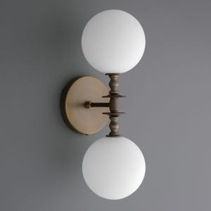 May include: A wall-mounted light fixture with two spherical, white glass shades. The fixture has a bronze-colored metal base and decorative accents. The design is symmetrical, with the shades positioned above and below a central support.