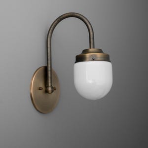May include: A vintage-style wall sconce with a curved, bronze-colored arm and a round, brass-colored wall plate. The light fixture features a white, rounded glass shade. The sconce is designed for indoor lighting.