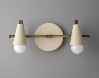 May include: Cream-coloured, two-light wall sconce with a circular backplate and a horizontal brass bar. Each light has a cone-shaped shade and a round, white bulb. The sconce has a minimalist, modern design.