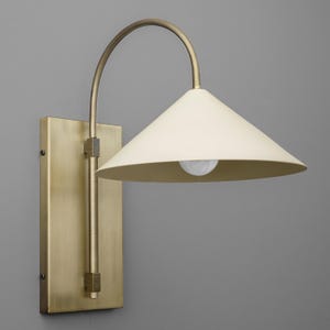 May include: A brass wall sconce with a cream-colored conical shade. The fixture has a rectangular backplate and a curved arm. The light bulb is visible inside the shade. The sconce is designed for indoor lighting.