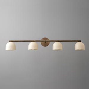 May include: A wall-mounted light fixture featuring a horizontal bronze-toned metal bar and a circular wall plate. Four dome-shaped, off-white shades are evenly spaced along the bar, each pointing downward to provide focused illumination.