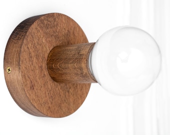 May include: A wall-mounted light fixture featuring a circular dark brown wooden base and a cylindrical wooden socket. A single round white glass light bulb is inserted into the socket. A small gold-colored screw is visible on the side of the base.