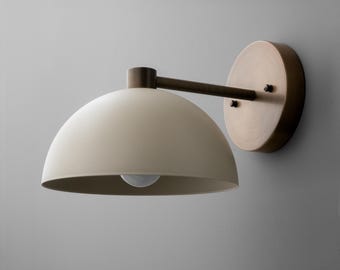 May include: Cream-colored wall sconce with a semi-circular shade and a brass-toned arm and mounting plate. The light fixture has a simple, modern design, with a visible lightbulb. The sconce is mounted on a gray wall.