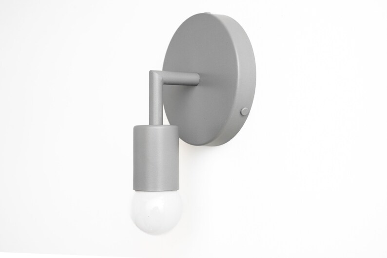 Puede incluir: Una l&aacute;mpara de pared plateada con una placa trasera redonda y una luminaria cil&iacute;ndrica. La luminaria tiene una bombilla de vidrio transparente.