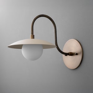 May include: A wall-mounted light fixture with a cream-colored shade and a white globe bulb. The fixture has a curved, dark bronze arm and a circular, brass-toned base. The design is minimalist and modern.